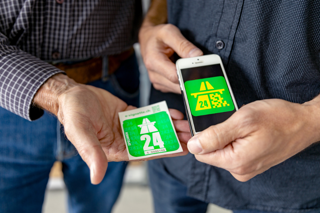 Klebevignette 2024 und Smartphone mit dem Logo der E-Vignette 2024 nebeneinander. Beide sind grün mit dem Autobahnzeichen und der Zahl 24.