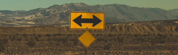 A steppe-like landscape with a yellow traffic sign in the foreground. On the sign are two arrows pointing in opposite directions.